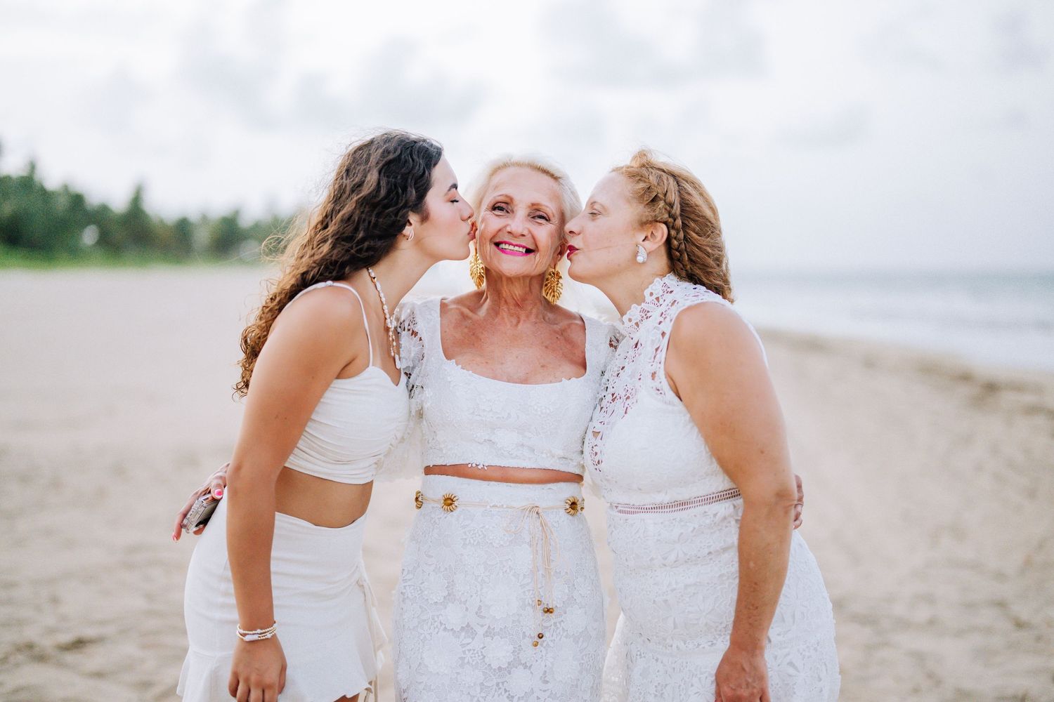 Three women sharing intimate moment on beach in white lace dresses.