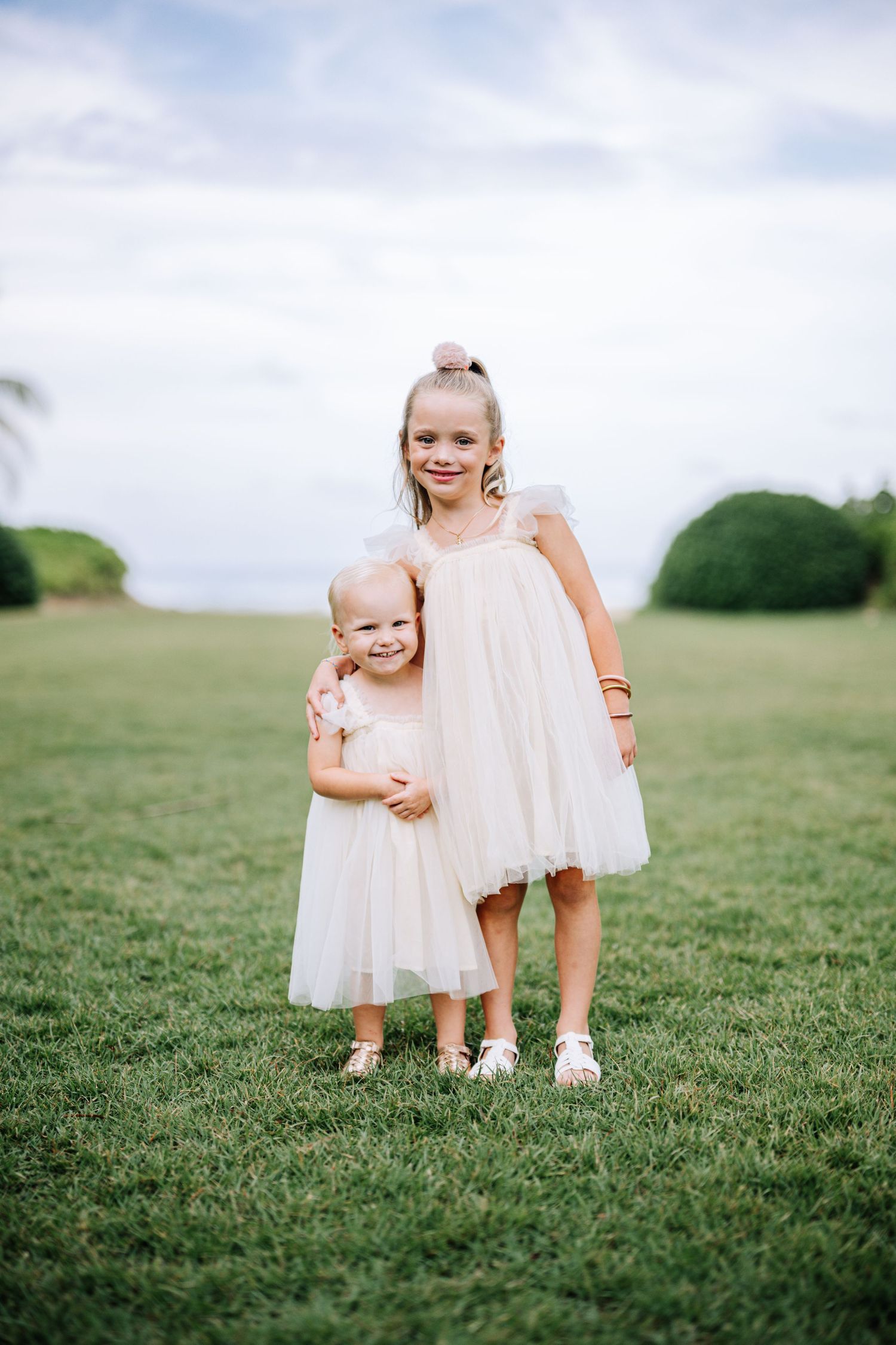 Two young children in light pink dresses standing together on grassy hill.