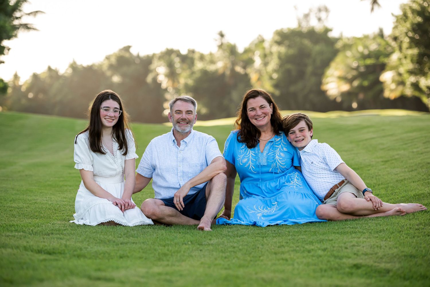 A family of four sits together on a grassy hill during golden hour at sunset.