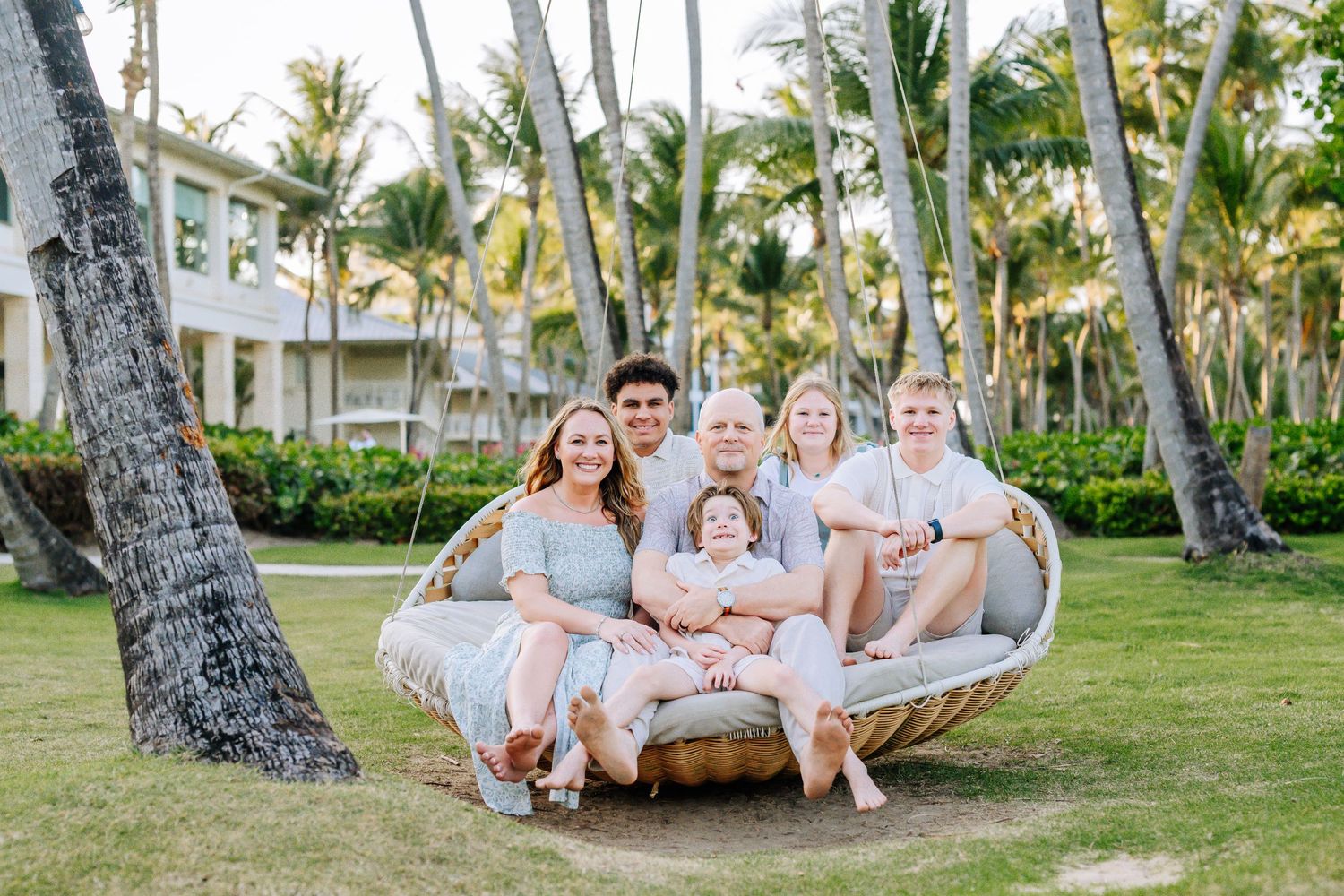 Family relaxing together in large outdoor hanging swing chair surrounded by palm trees.