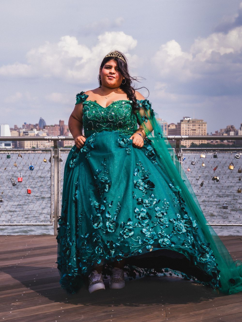 A stunning emerald green ball gown with floral appliques photographed against a waterfront cityscape background.