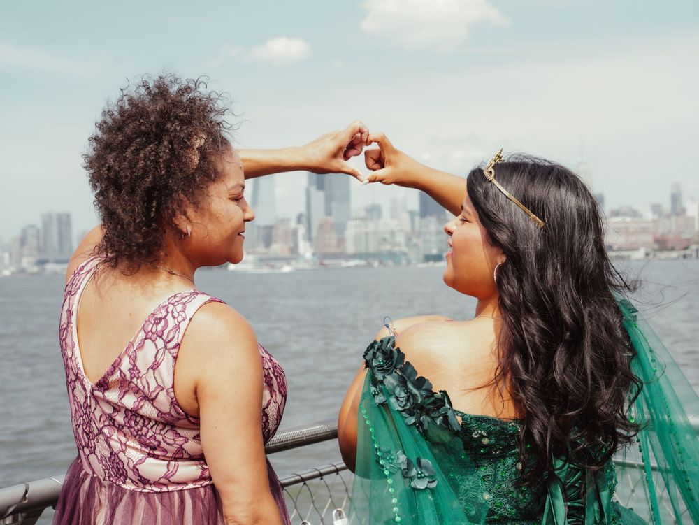 Two friends pose making a heart shape with their hands against a city waterfront backdrop.