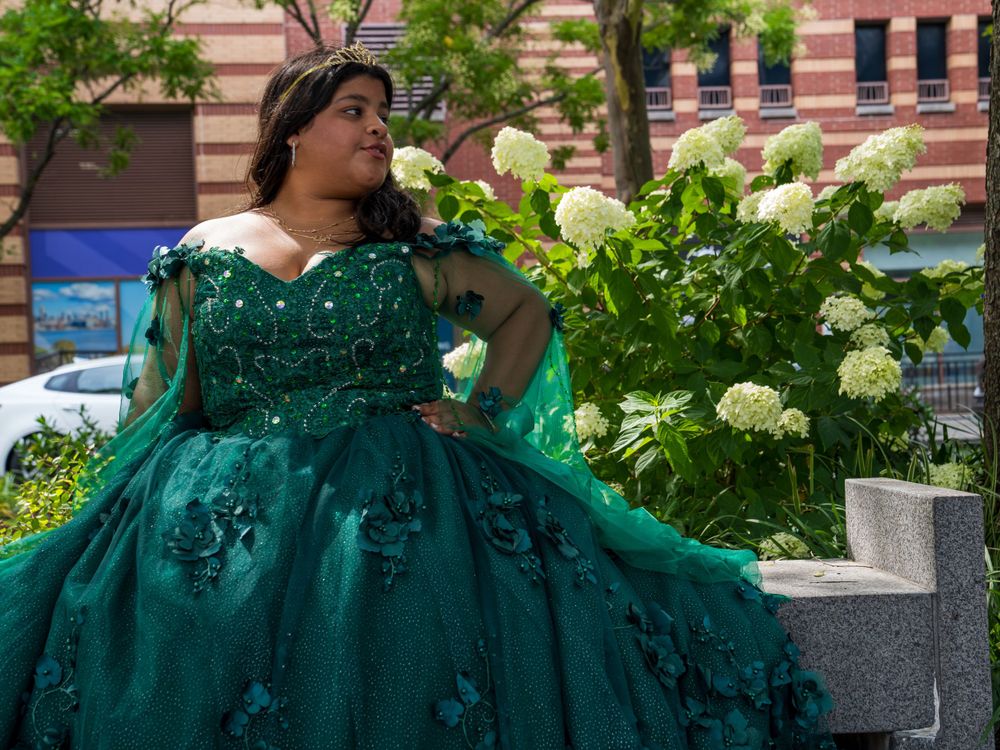 An ornate emerald green ball gown showcased against blooming white hydrangea bushes in an urban setting.