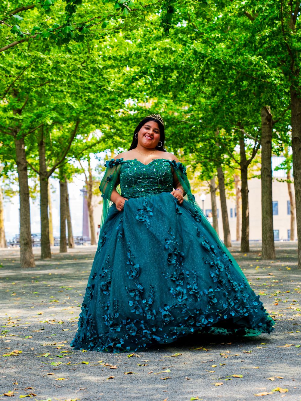 A flowing emerald green ball gown with floral details photographed in a tree-lined outdoor setting.