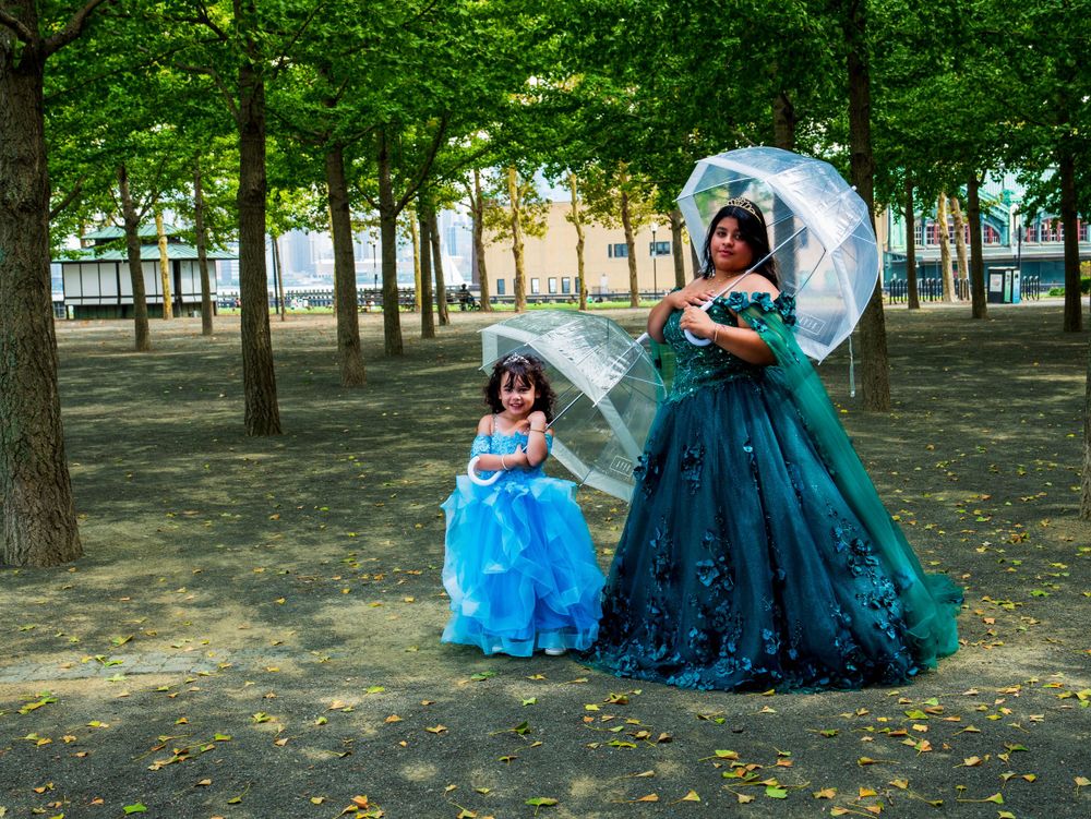 Two people in formal dresses holding parasols pose among trees in a park setting.
