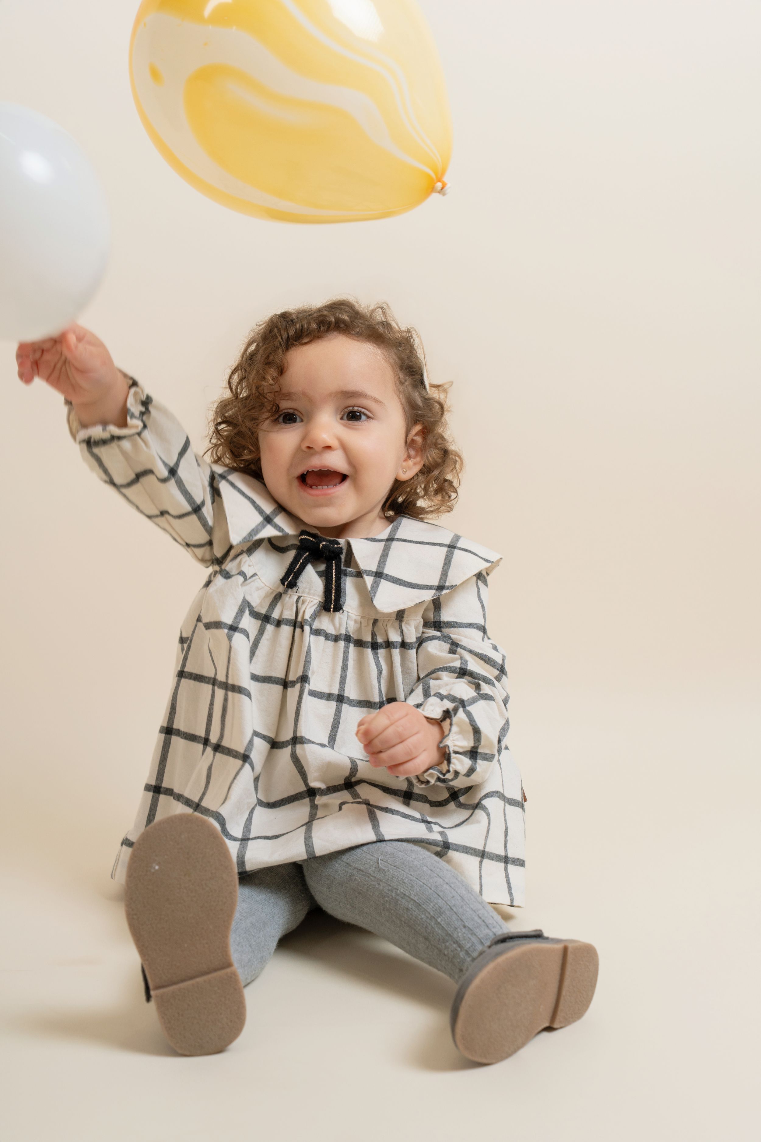 A smiling toddler girl in a checkered dress sitting on the floor reaching up toward colorful balloons.