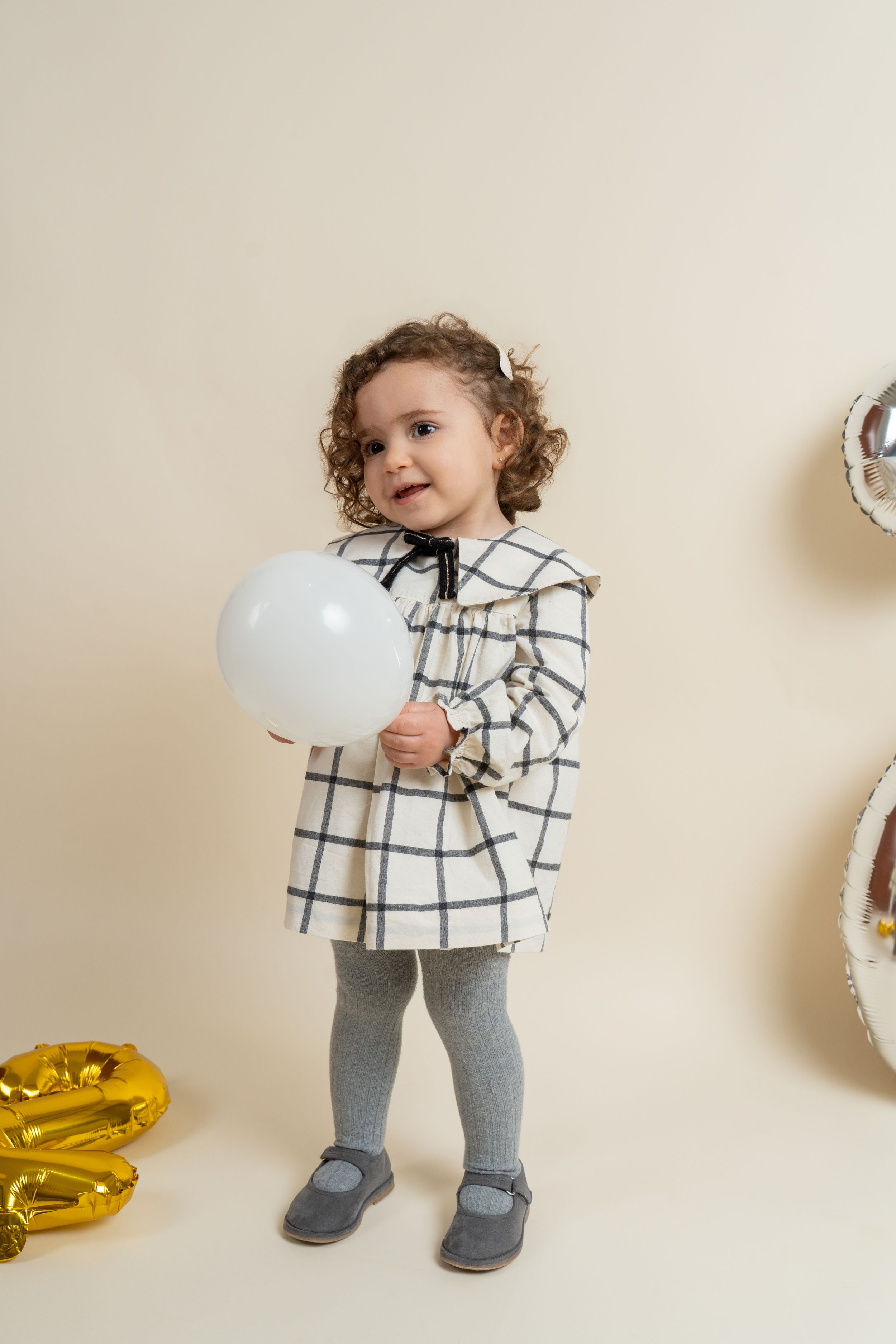 A curly-haired toddler girl in a plaid dress holding a white balloon surrounded by gold and silver balloons.