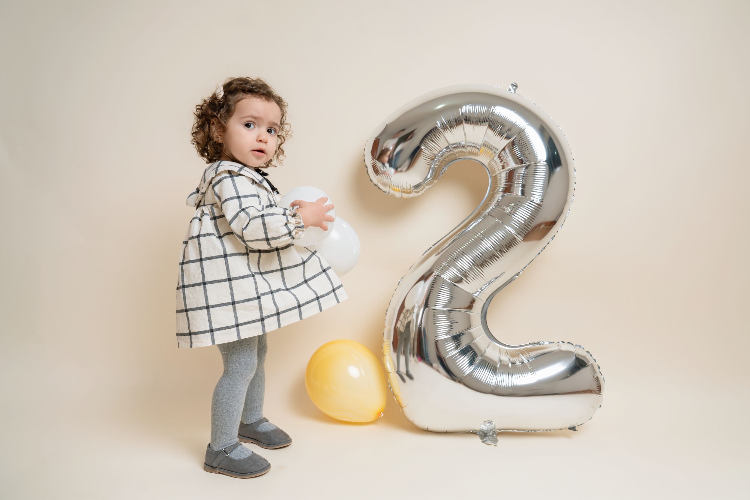 A cute toddler girl in a plaid dress standing beside a large silver number 2 balloon for her birthday.