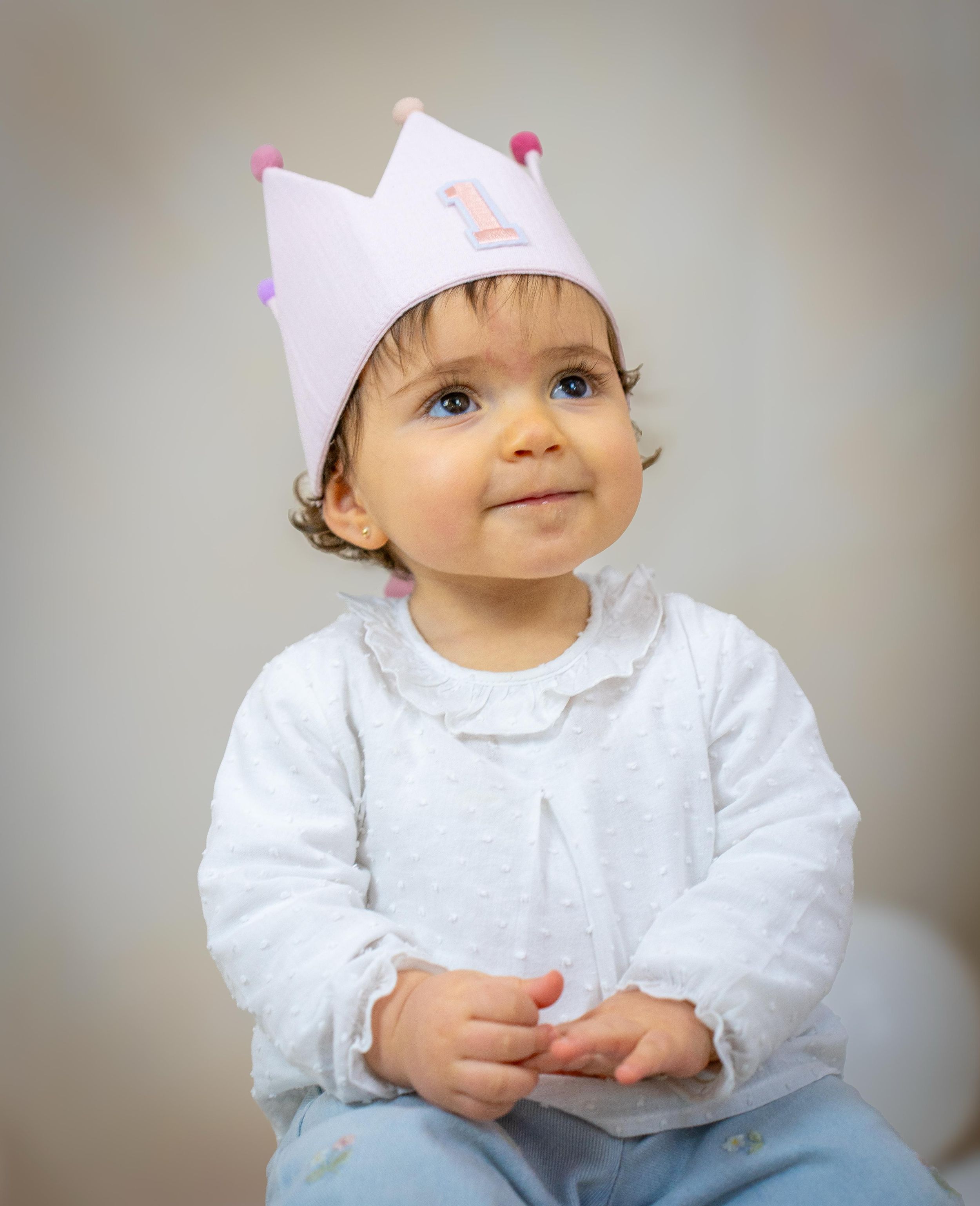 Adorable baby girl wearing pink number 1 birthday crown smiles sweetly in white outfit for first birthday.