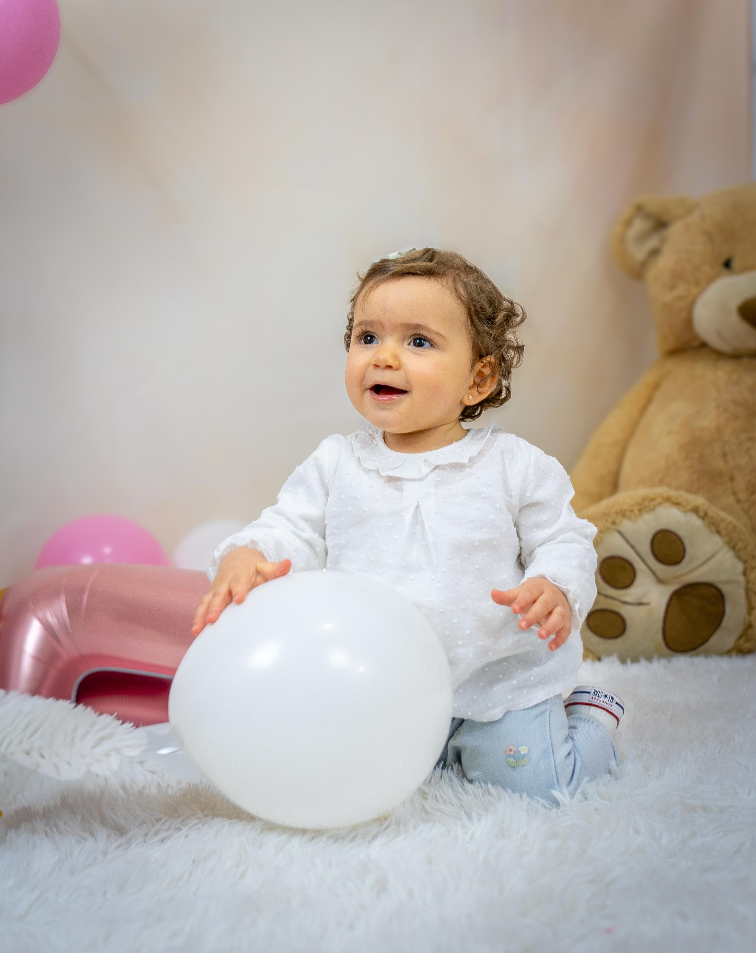 Baby girl in white dress plays with white balloon surrounded by pink balloons and teddy bear on white fluffy rug.