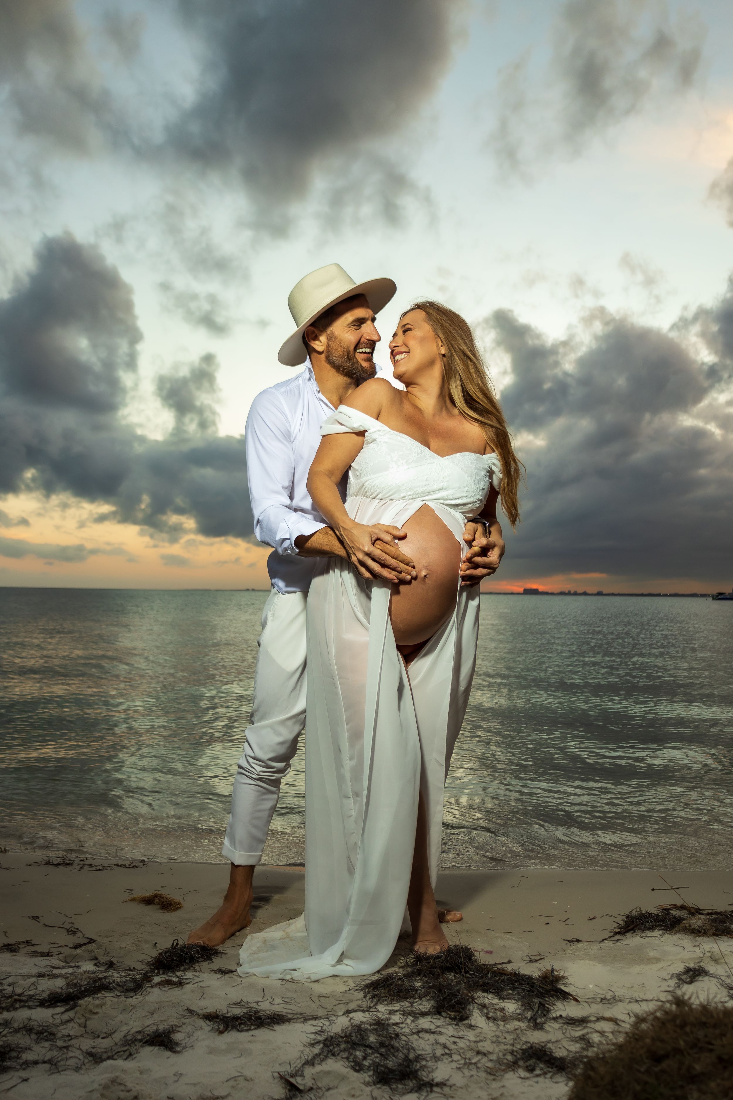 Elegant beach maternity photoshoot at sunset with flowing white dress.