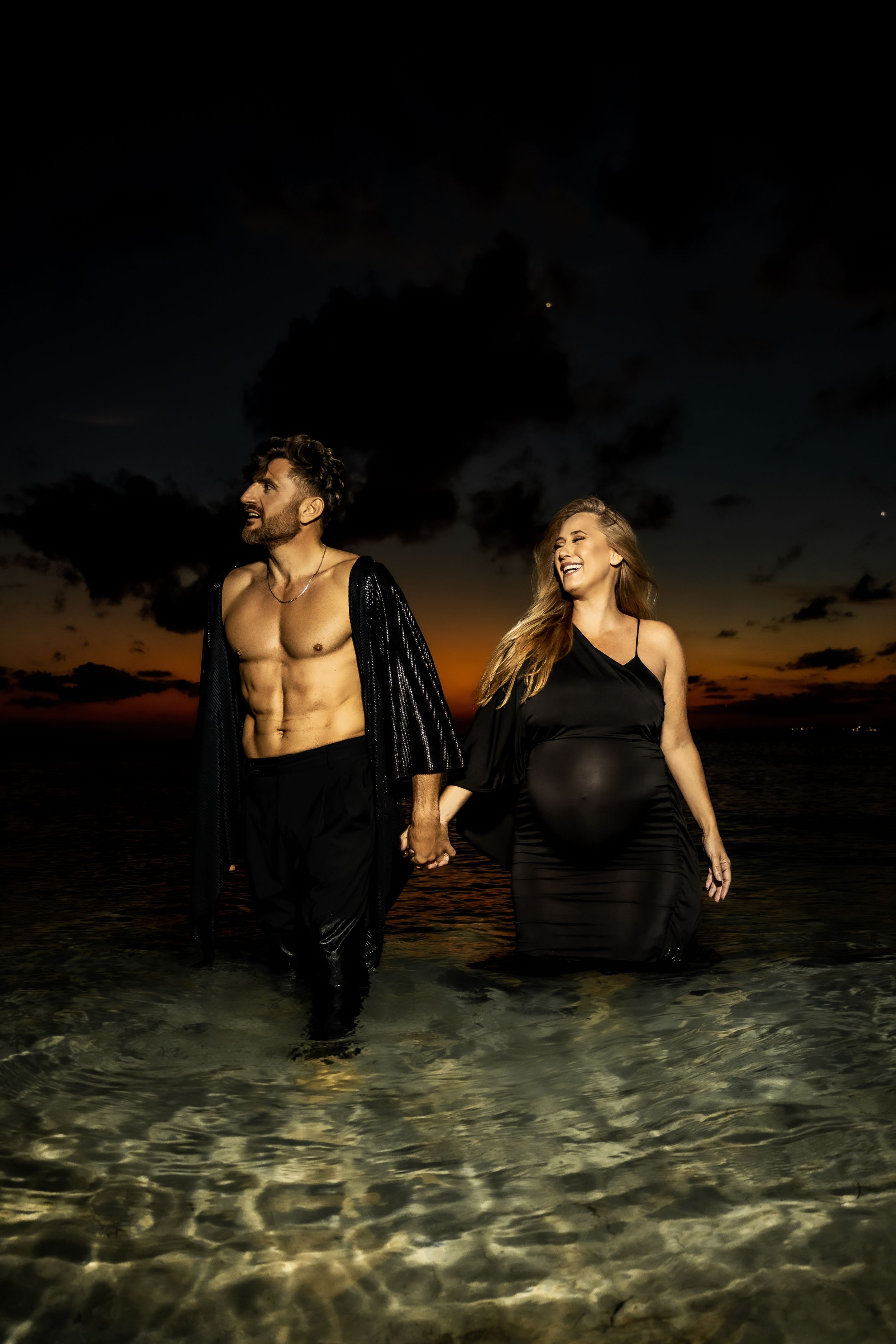 A romantic maternity photoshoot at sunset with a couple standing in shallow ocean water.