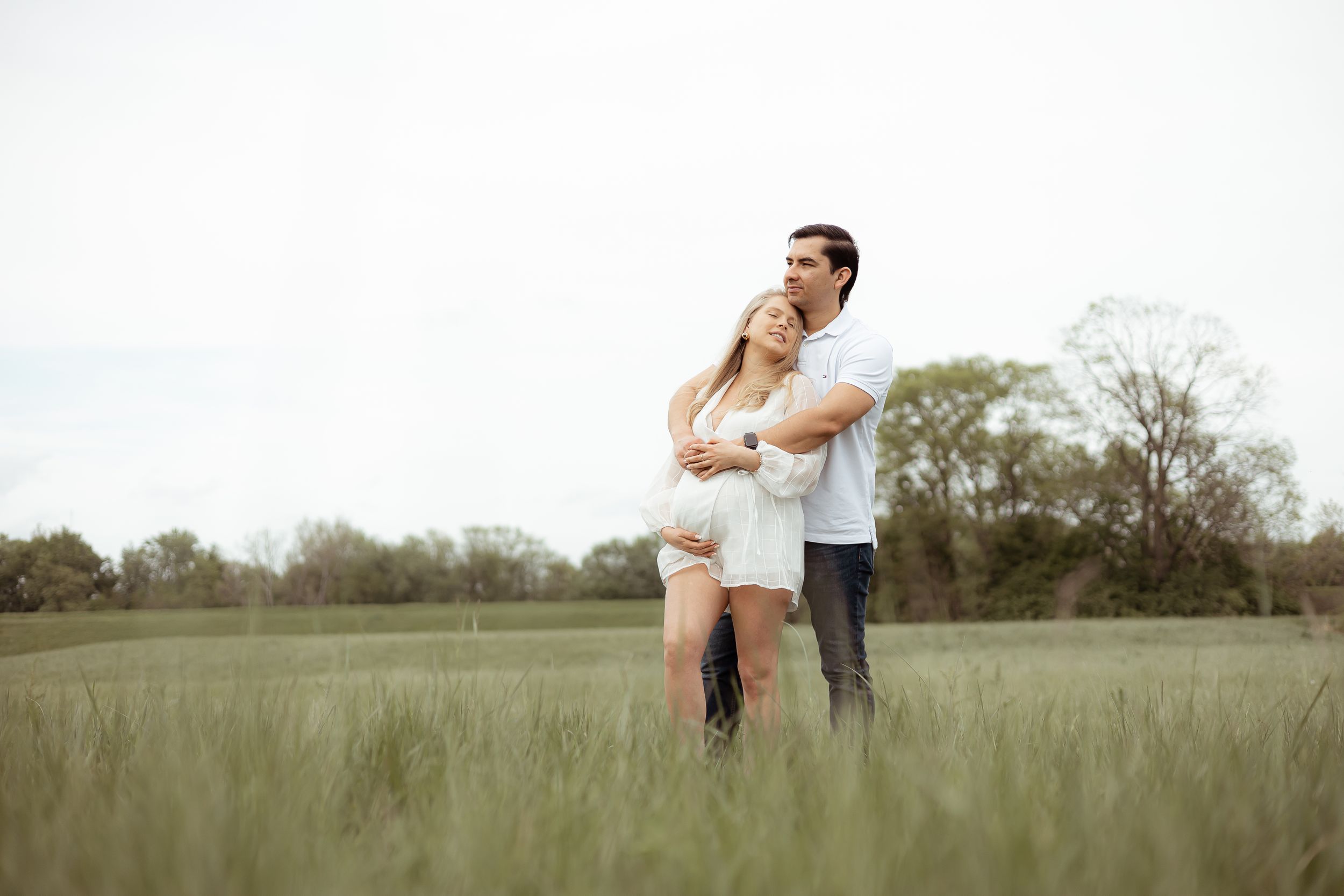 Maternity couple standing together in tall grass field.