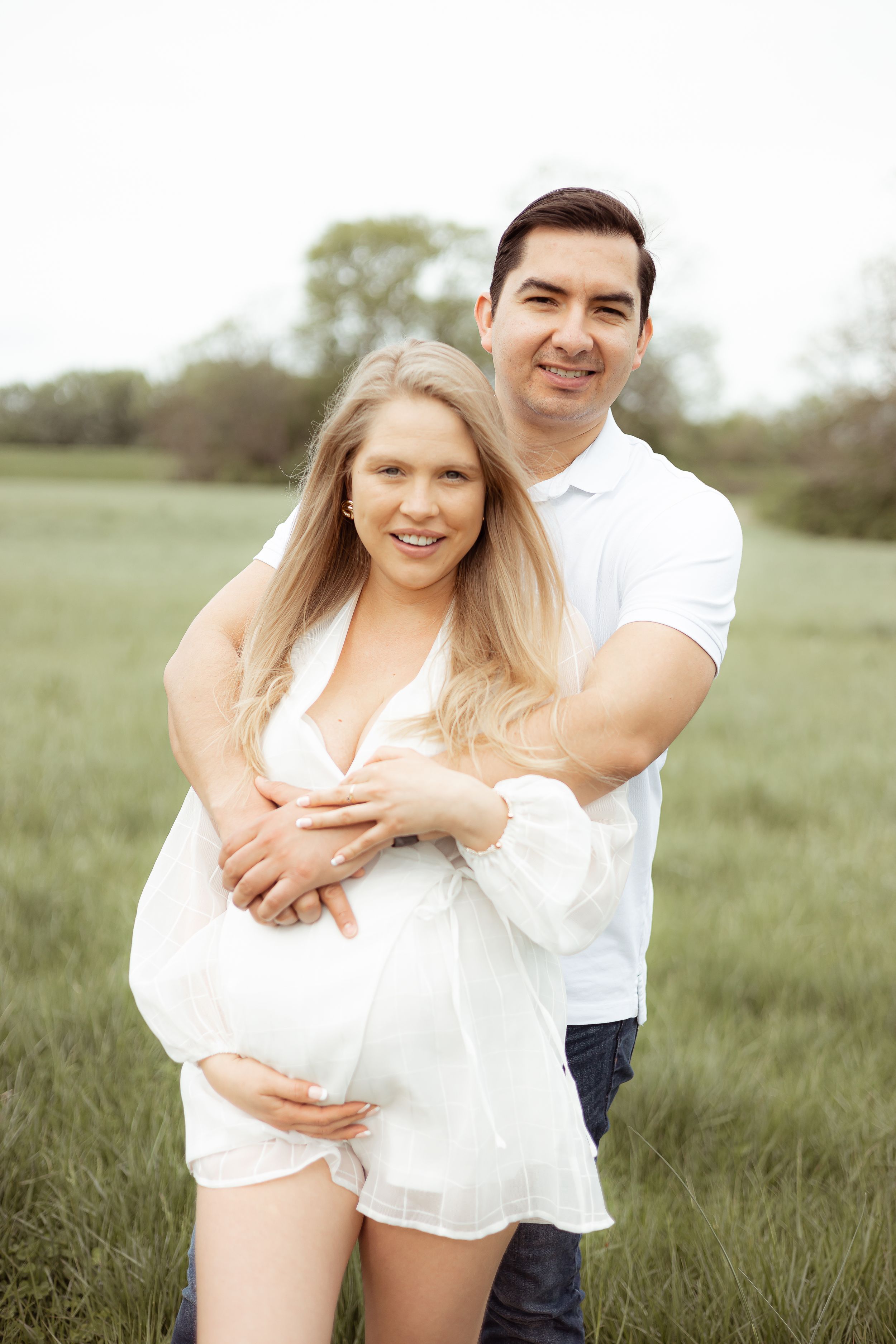 Expecting couple embracing in sunny green field wearing white clothing.