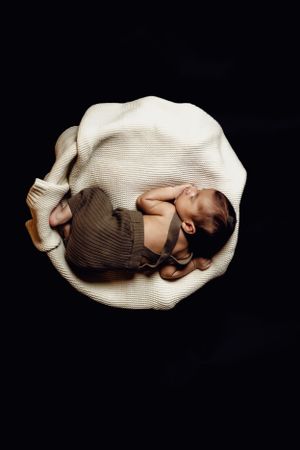 Newborn baby curled up on white blanket against black background in artistic overhead shot.