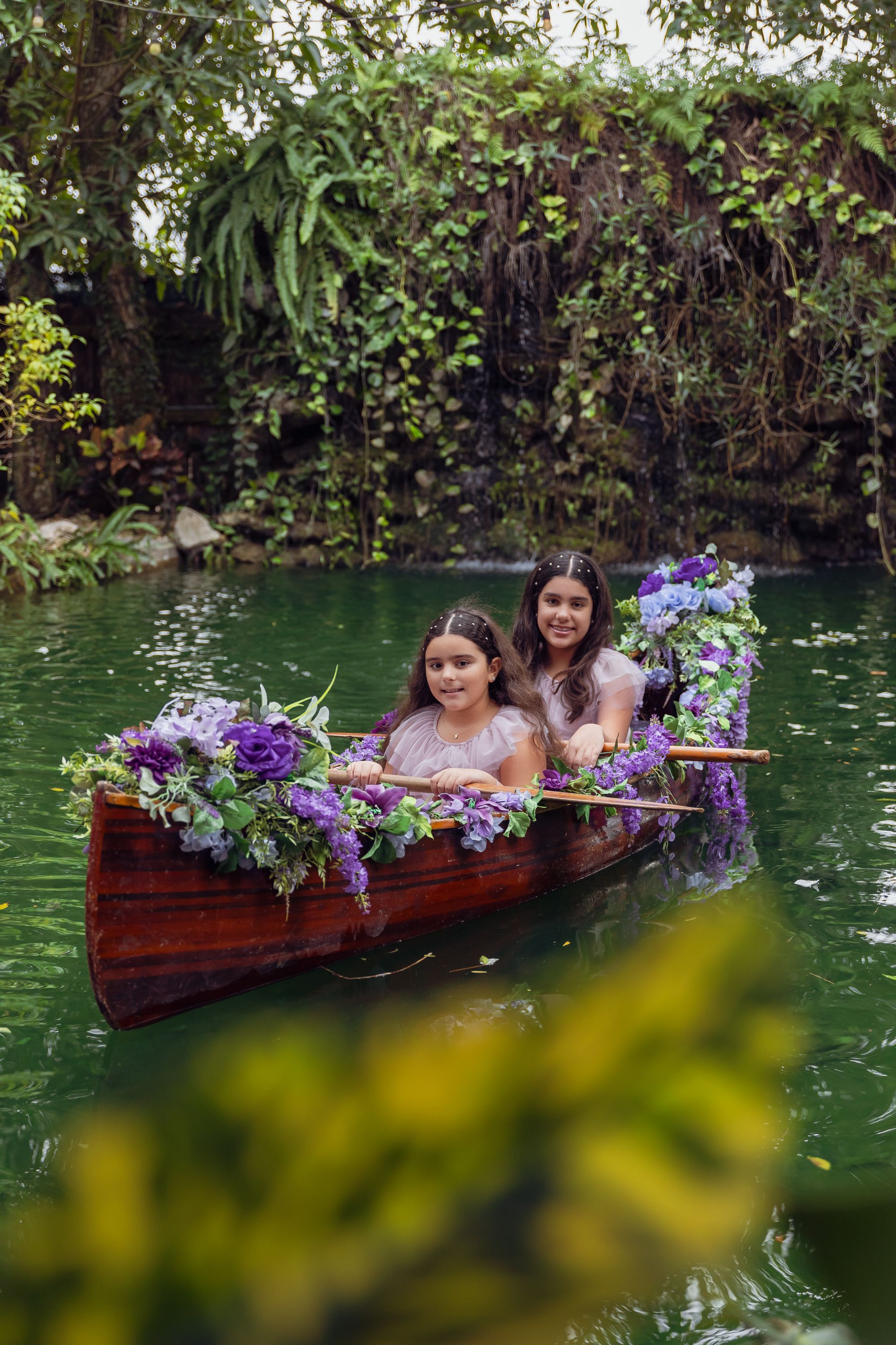 Two people in floral decorated wooden boat on green pond surrounded by hanging vines.