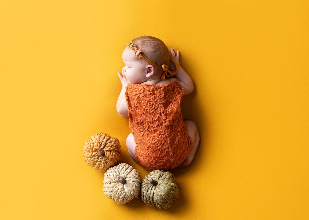 Baby wearing orange knit outfit lying on bright yellow background with decorative pumpkins.