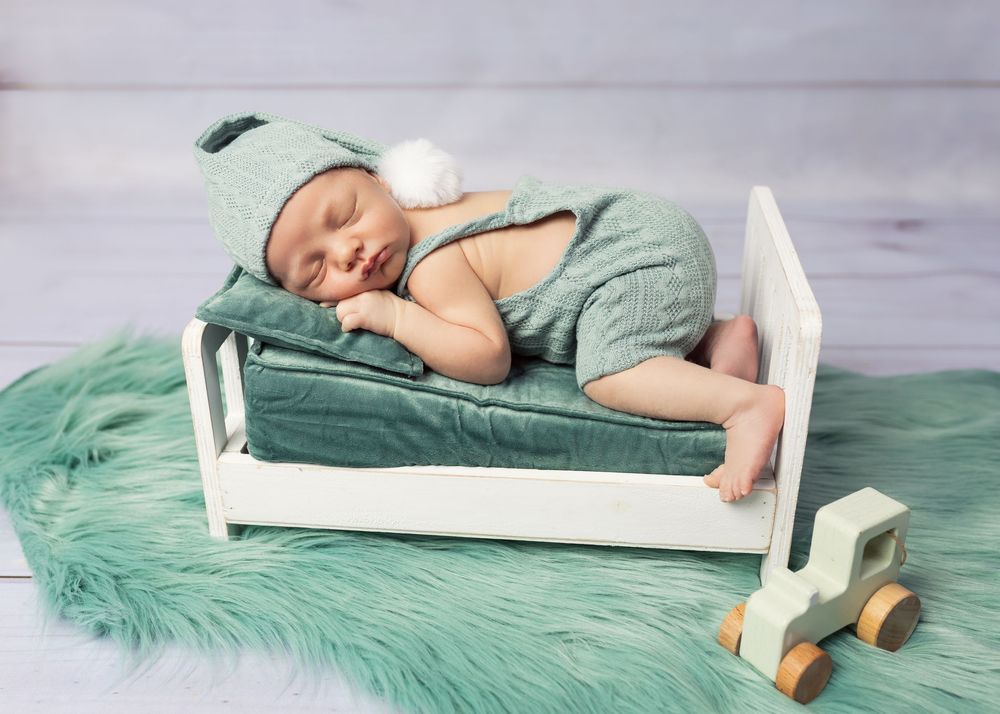 Baby sleeping on miniature white bed with mint green accessories and wooden toy car.