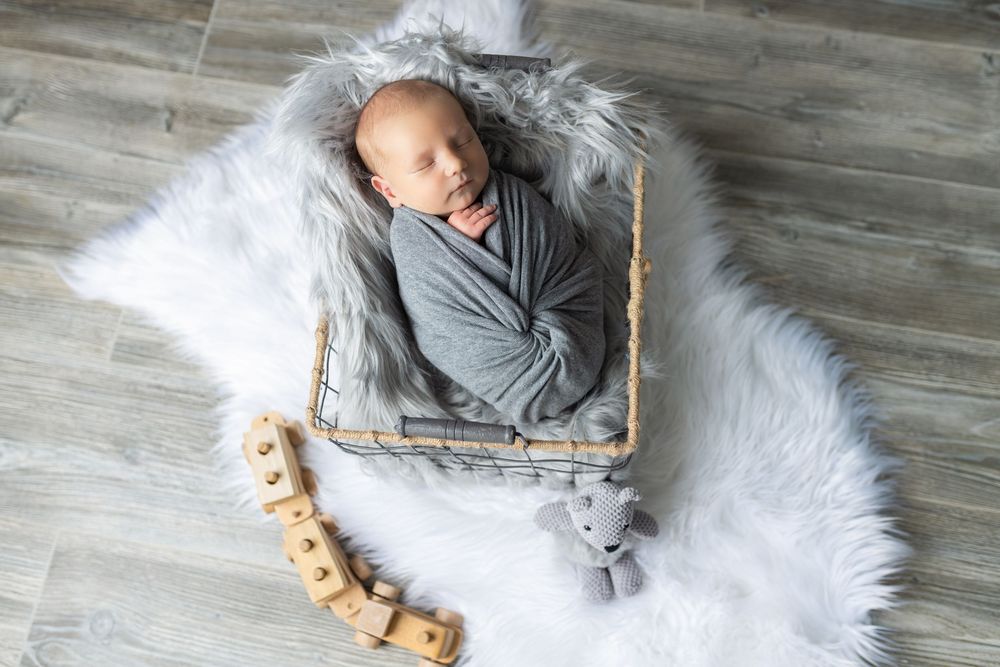 Overhead view of sleeping baby wrapped in gray swaddle in wooden basket on white fur.