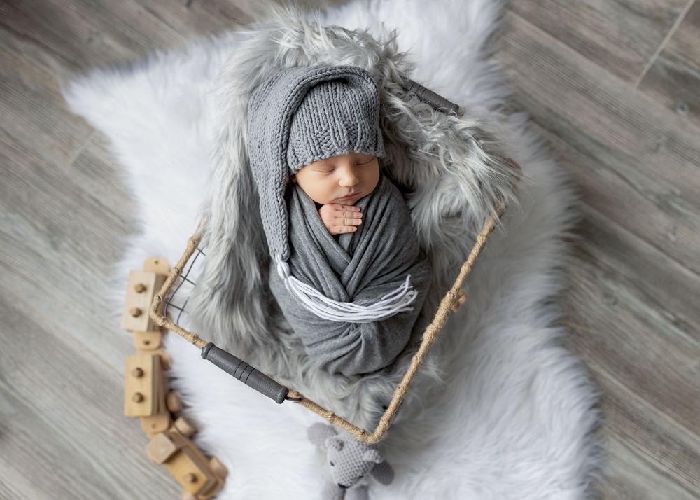 Newborn in gray knit hat and wrap sleeping in wooden basket with wooden toys.