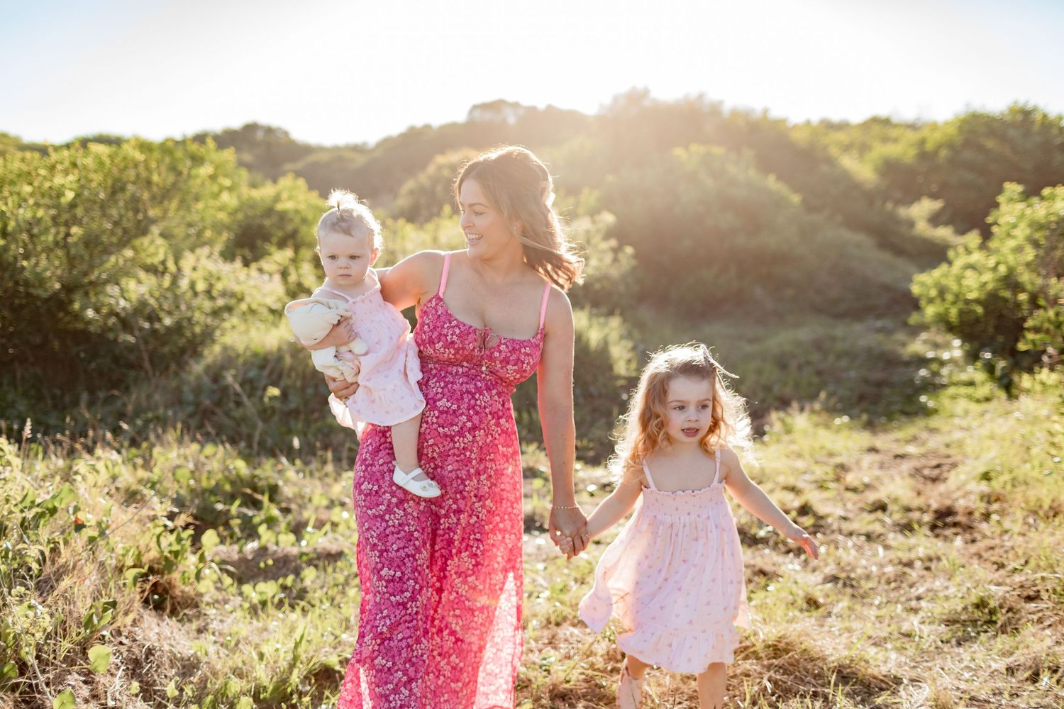 Family walks together through golden sunlit field wearing coordinating pink and white outfits.