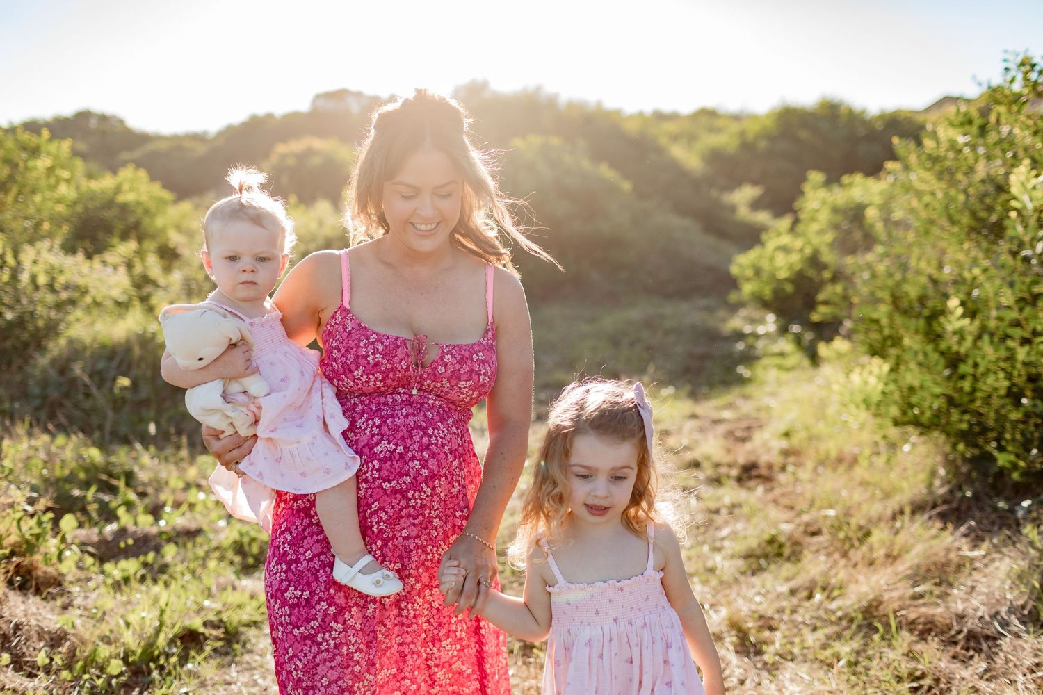 Three people stroll through mountainside meadow at sunset in matching pink and white attire.