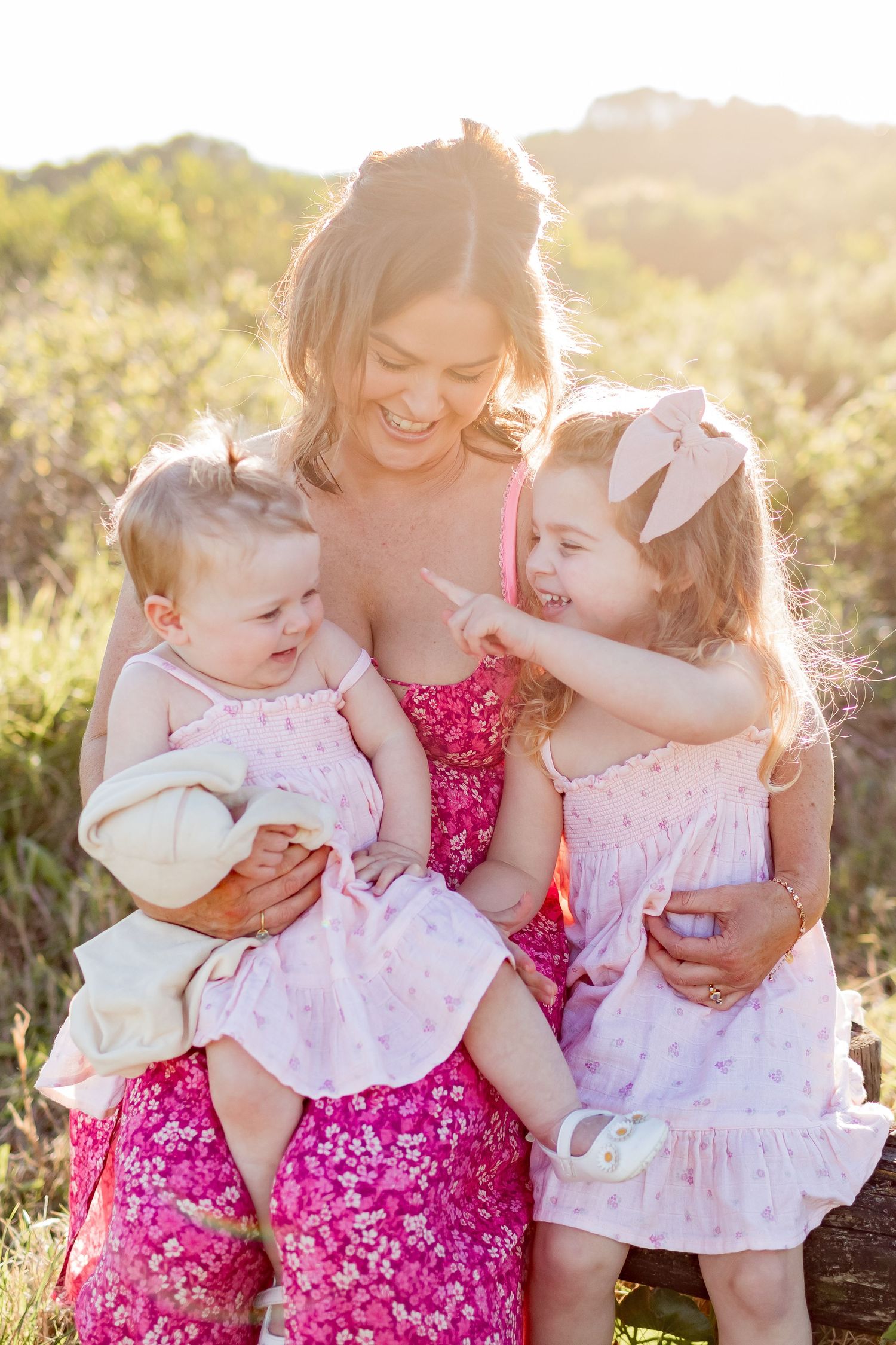 Joyful moment shared between three people wearing pink and white dresses in sunny field.