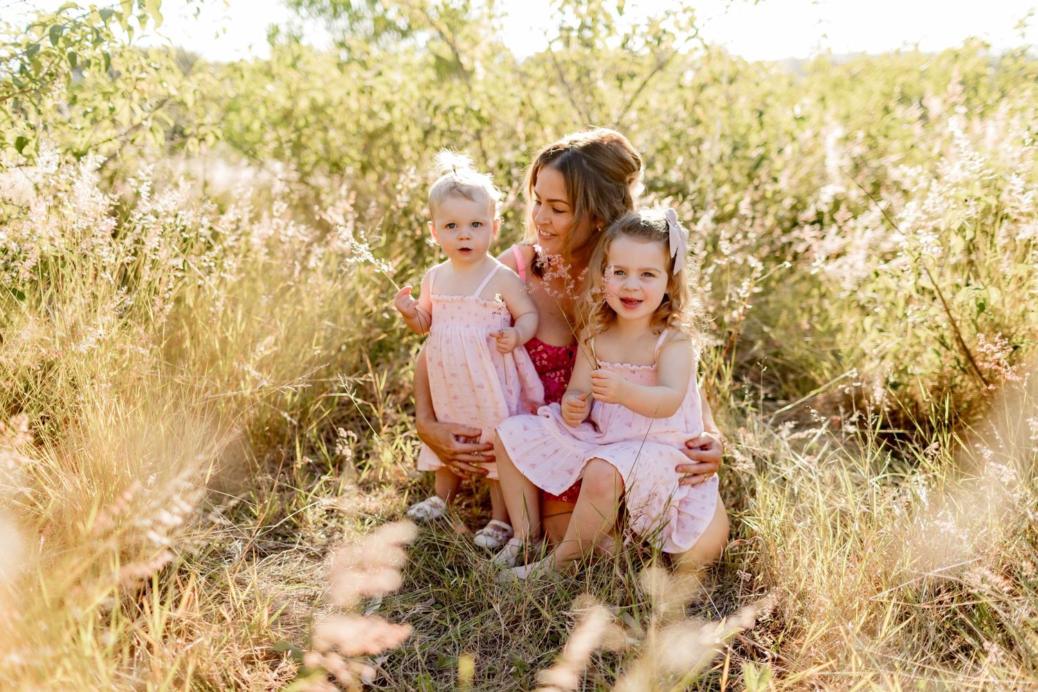 Three people in pink and white dresses share intimate moment in golden sunlit field.