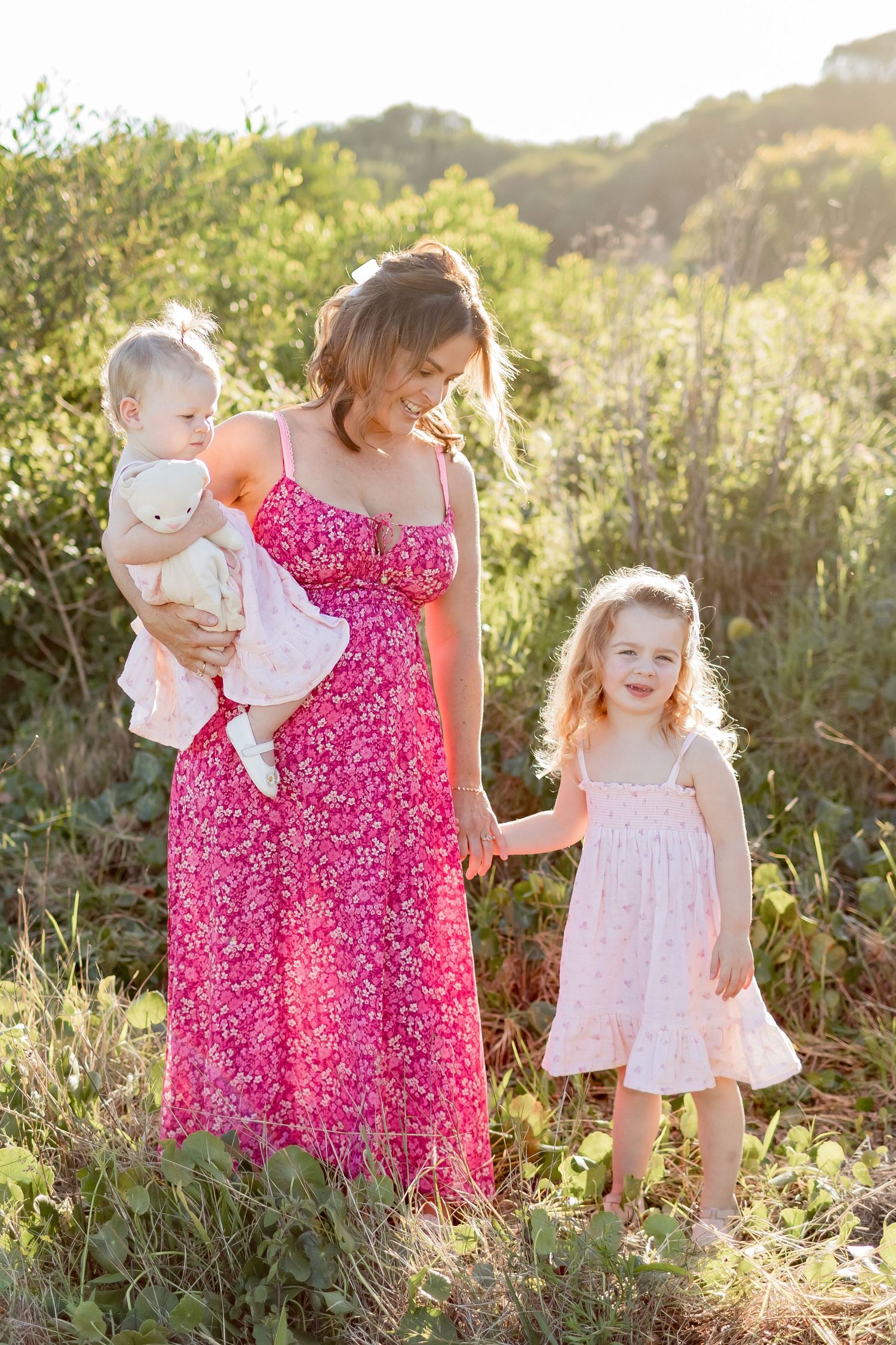 Three people share a tender moment in a sunlit field wearing pink and white dresses.