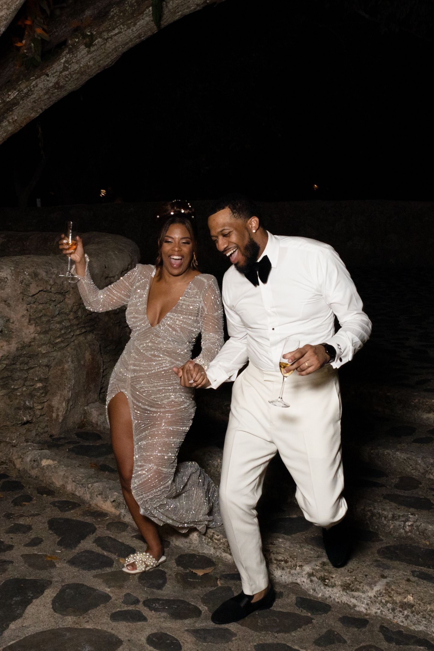 A couple in elegant attire dances together in a stone archway, wearing a sparkly gown and white tuxedo.