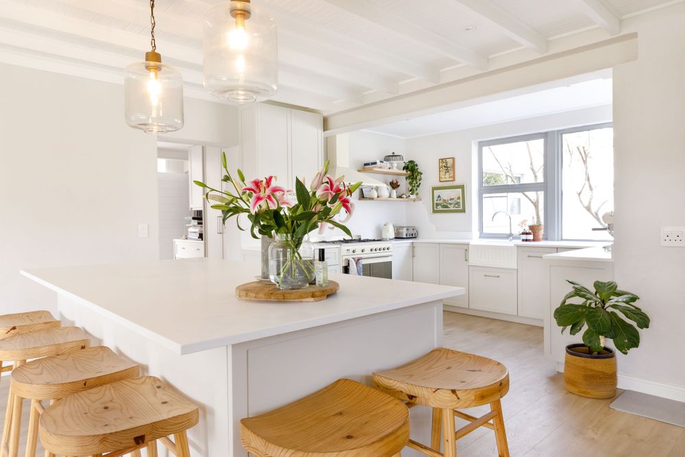 White coastal dining area with wooden bar stools and fresh floral arrangement on counter.