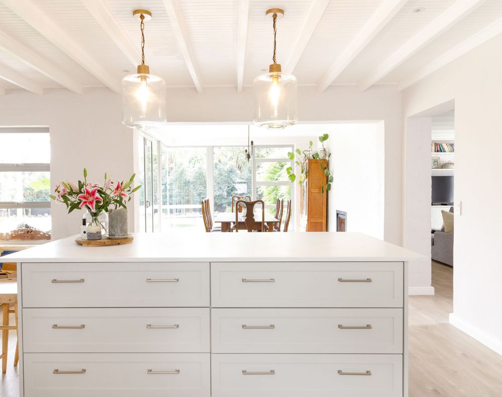 White kitchen island with brass pendant lights and drawers against bright window backdrop.