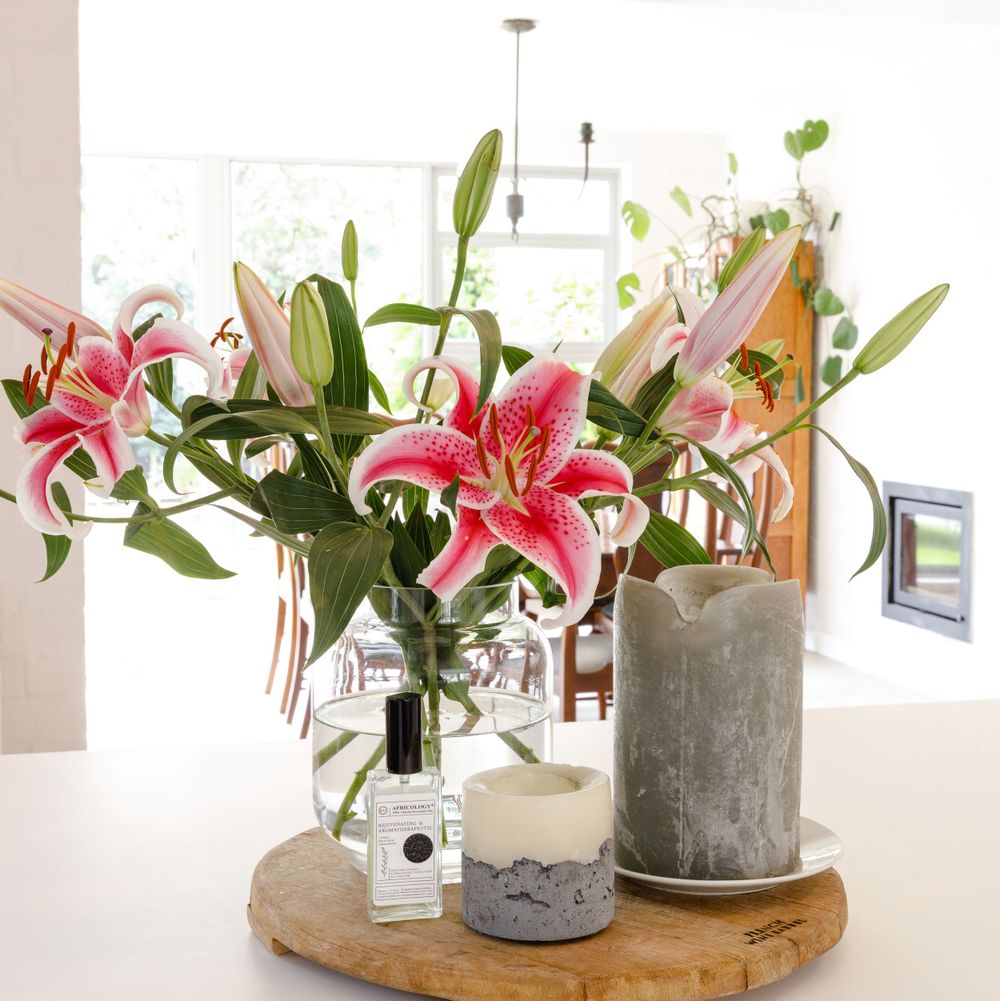 Pink stargazer lilies in glass vase with decorative candles on wooden board.