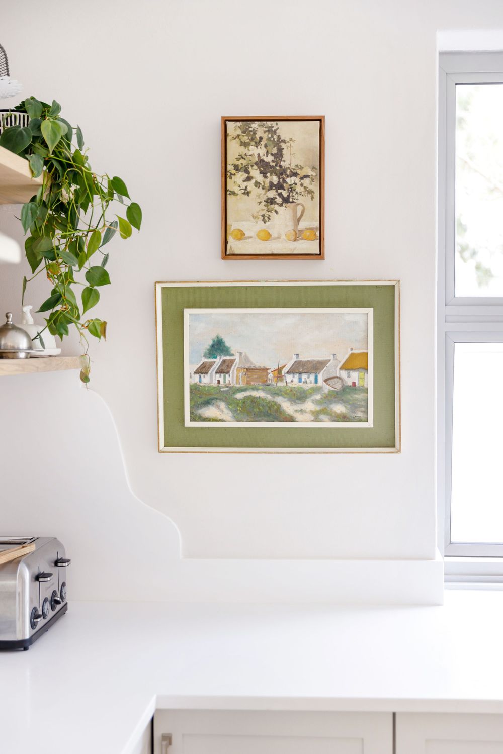 Kitchen wall decorated with framed artwork featuring houses and trailing potted plant.
