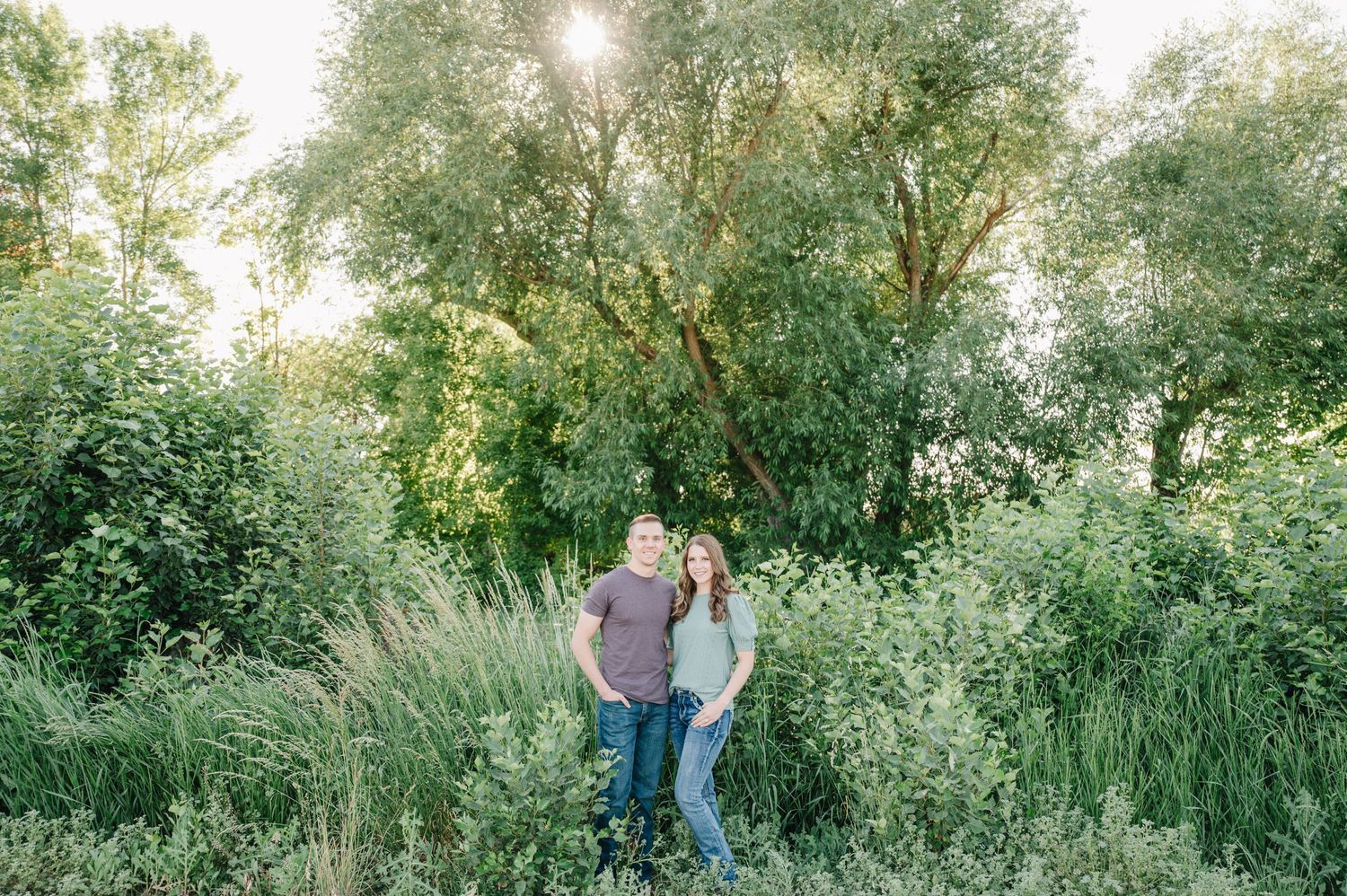 Couple standing together in meadow with backlit trees during sunset.