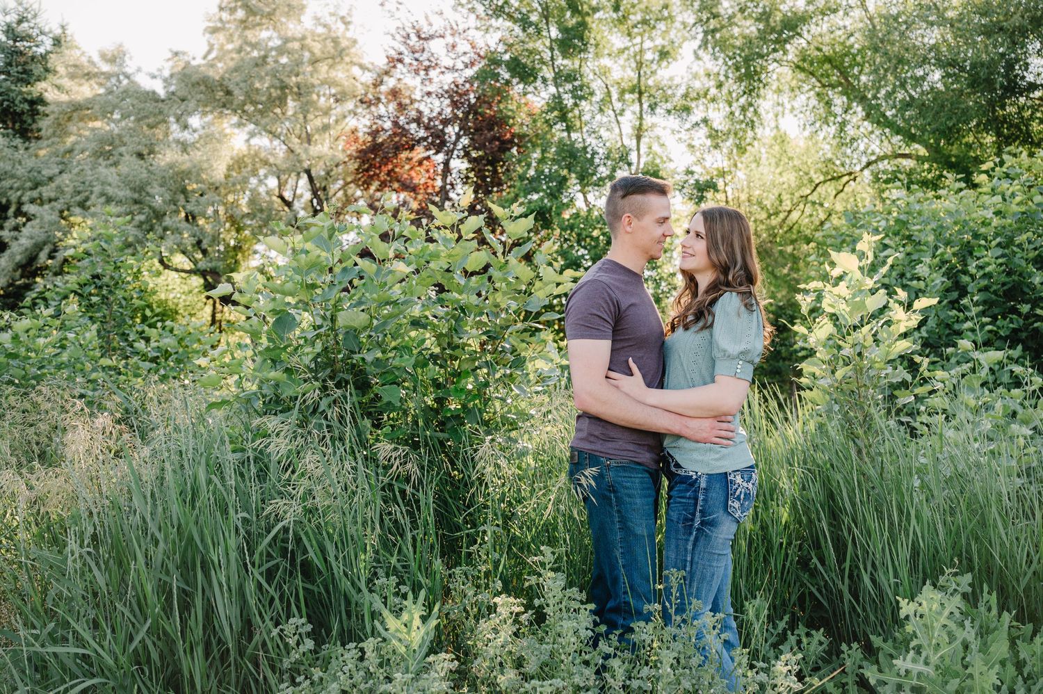 Couple embracing in tall grass with sunlit trees in background.