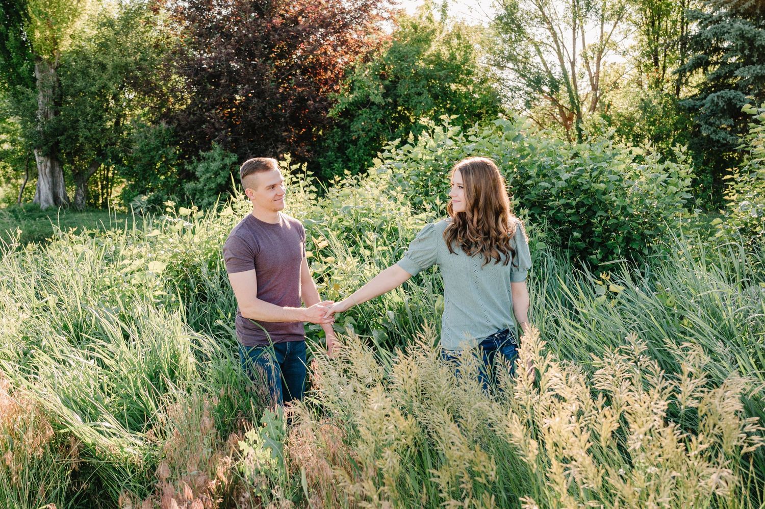 Couple holding hands while standing in wild grass field with trees behind them.