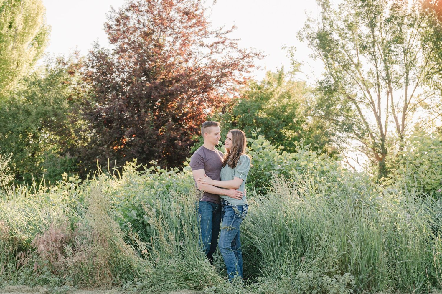 Couple sharing intimate moment in tall grass with sun-dappled trees behind.