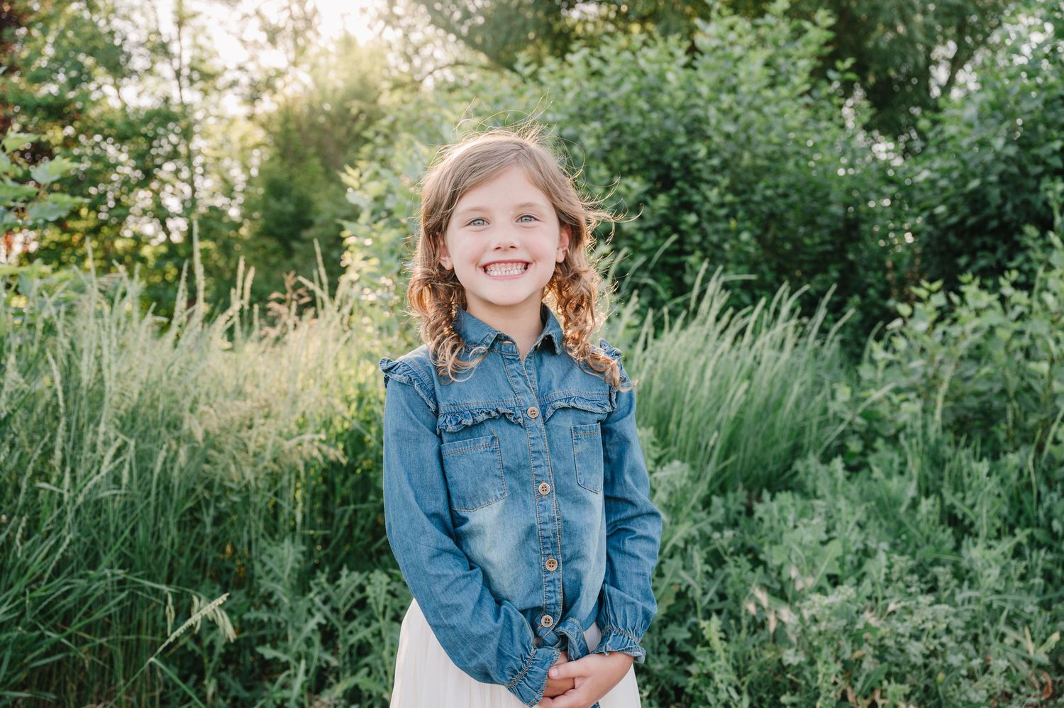 Young person wearing denim jacket smiles outdoors with green foliage in background.