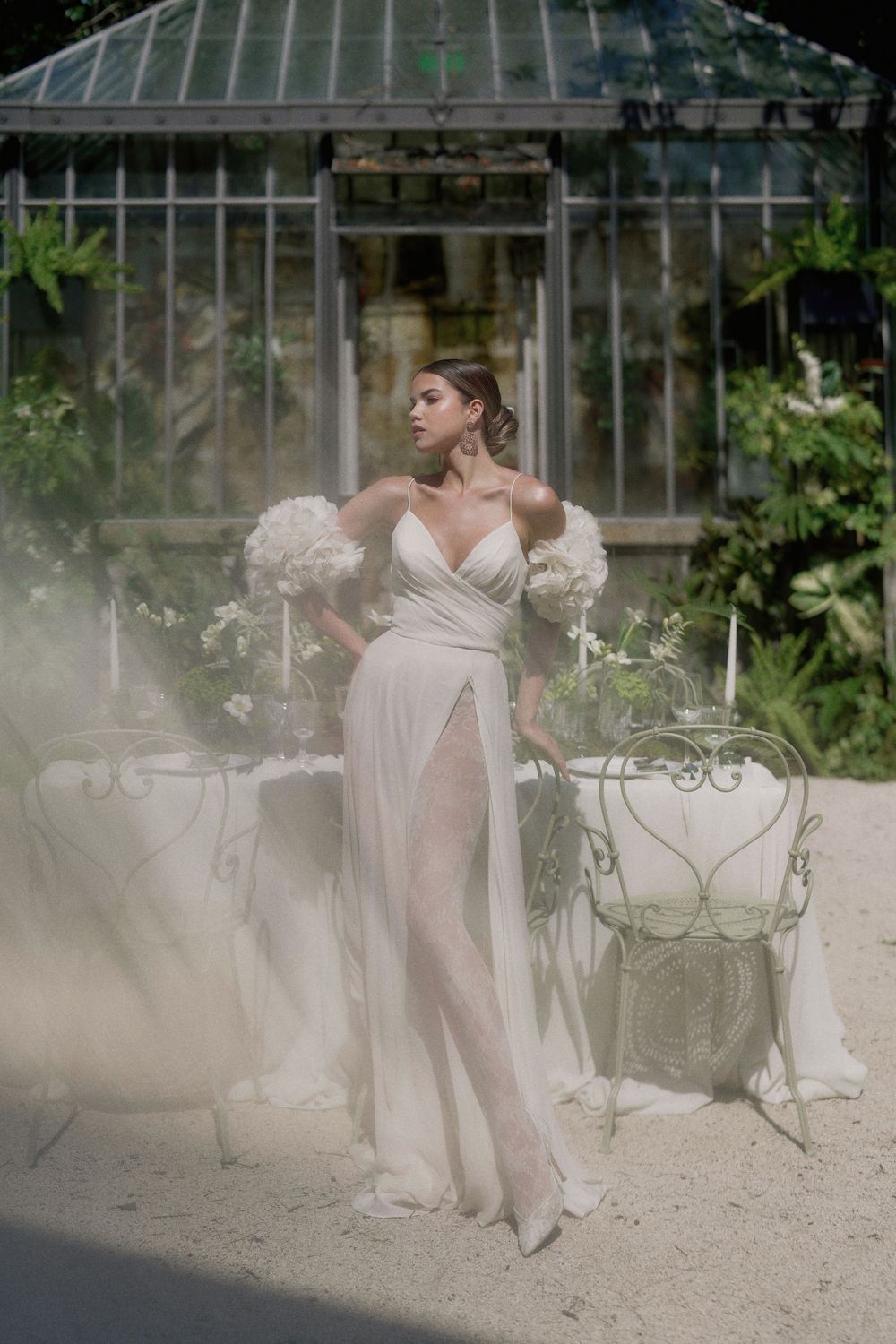 Ethereal bridal portrait featuring a white flowing gown with dramatic sleeves in a misty greenhouse setting.