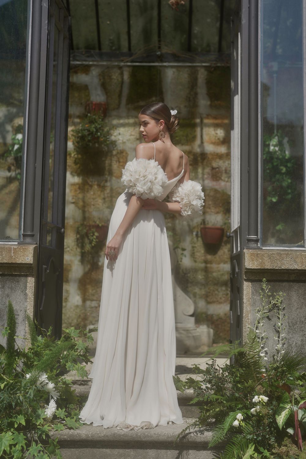 Elegant off-shoulder white wedding dress with flowy fabric photographed in front of a rustic greenhouse door surrounded by flowers.