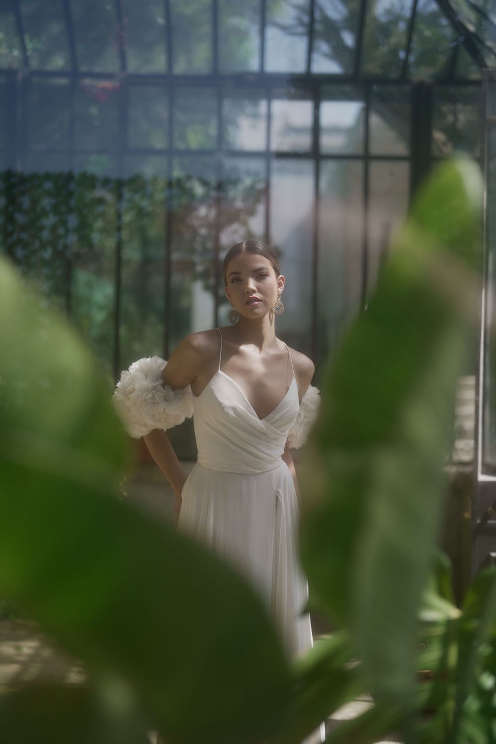 Elegant white wedding dress photographed through greenhouse glass and leaves in soft, dreamy lighting.