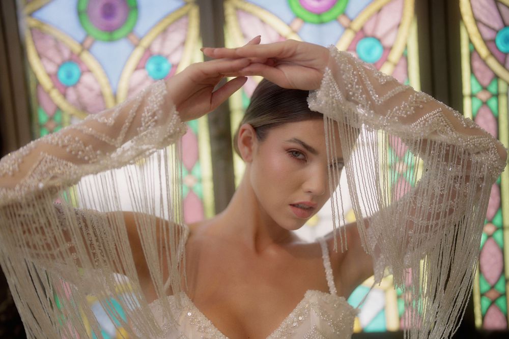 Ethereal figure in delicate lace sleeves poses against colorful stained glass window backdrop in artistic portrait.