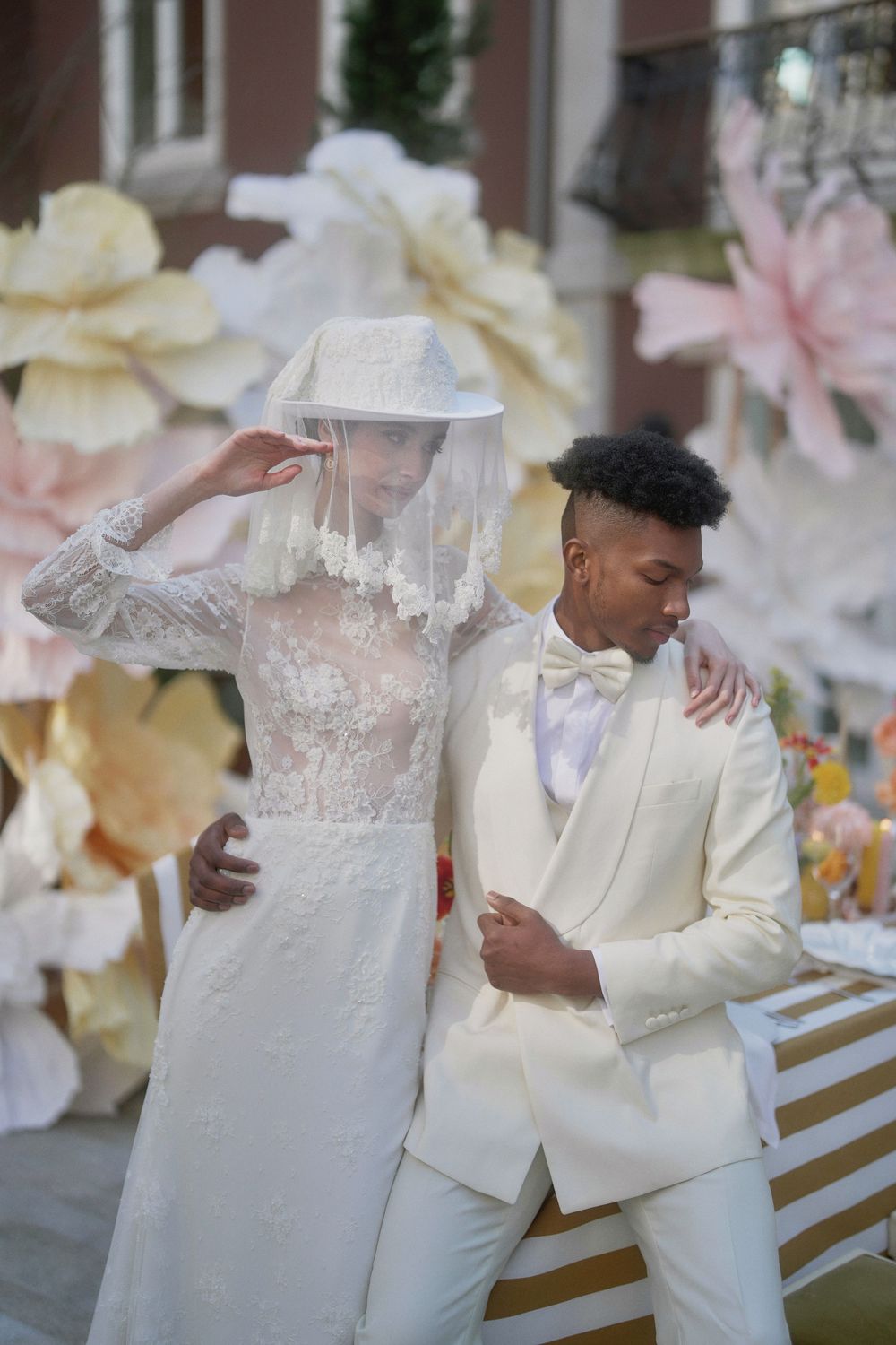 A newlywed couple wearing all white outfits pose together against a backdrop of large decorative flowers at their wedding.