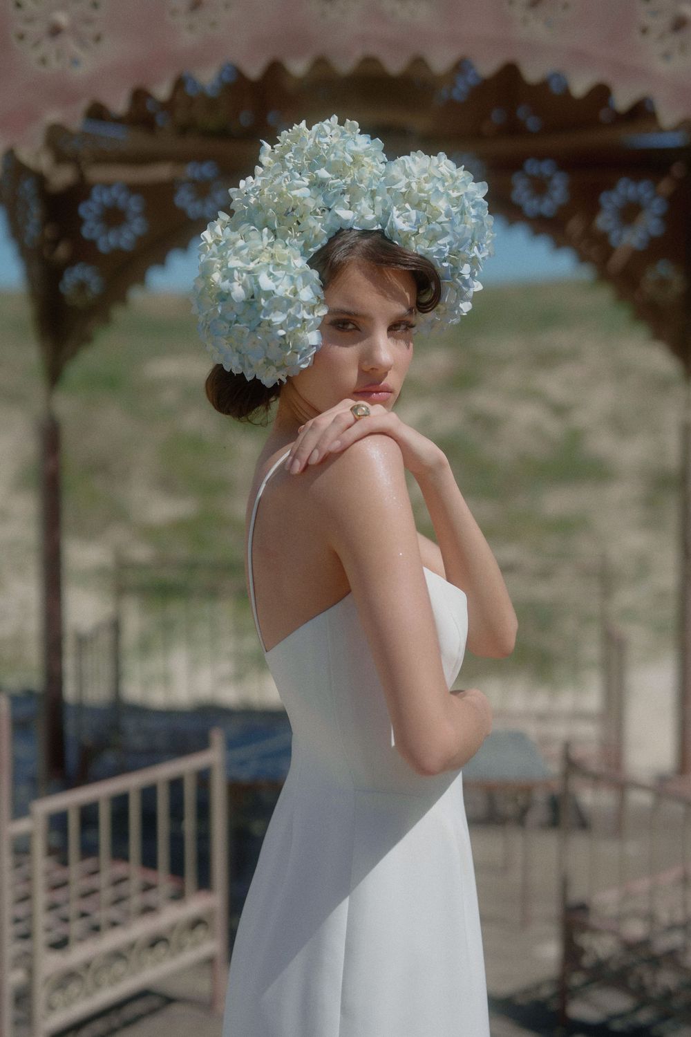 A bride in an elegant white dress poses under a gazebo while wearing a crown of delicate blue hydrangeas.