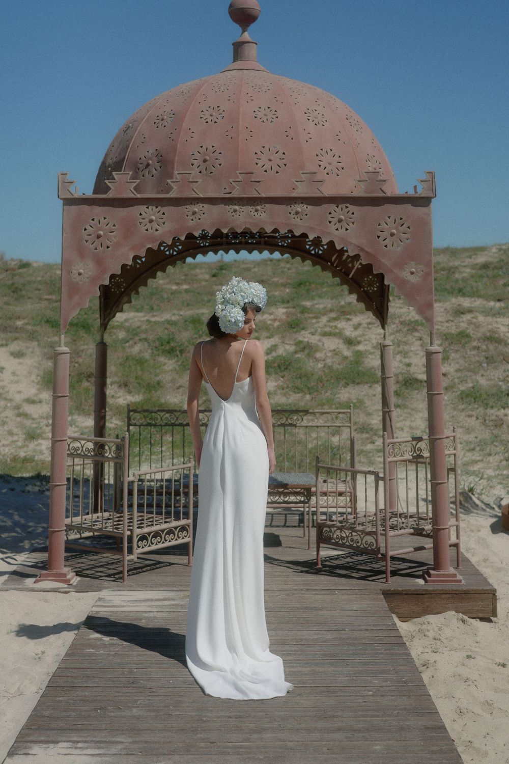 A white wedding dress photographed from behind under a pink domed gazebo at a beach location.