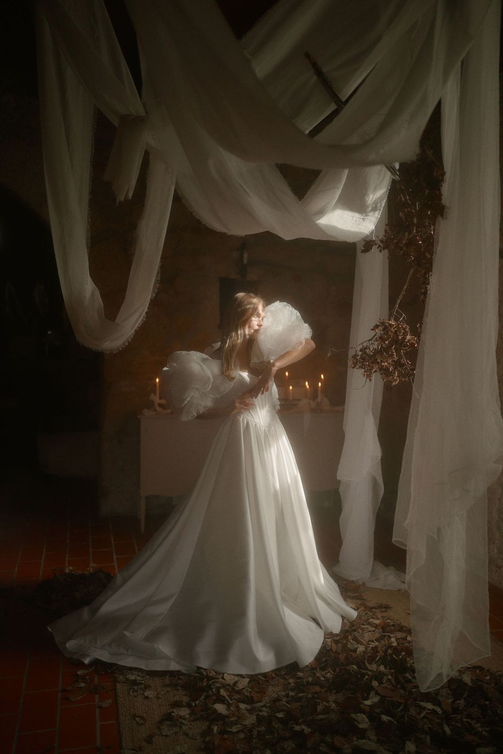 Ethereal wedding dress glows in candlelight under flowing white fabric drapes and scattered autumn leaves in a moody setting.