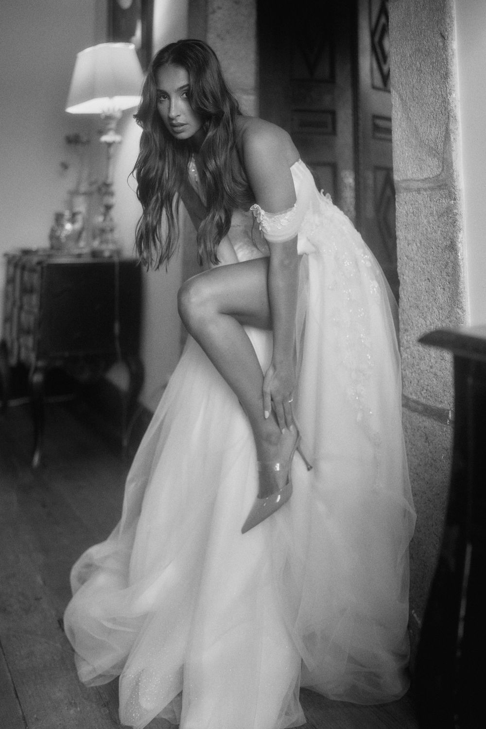 A dramatic black and white boudoir photo of someone in a flowing white dress against a dark interior setting.