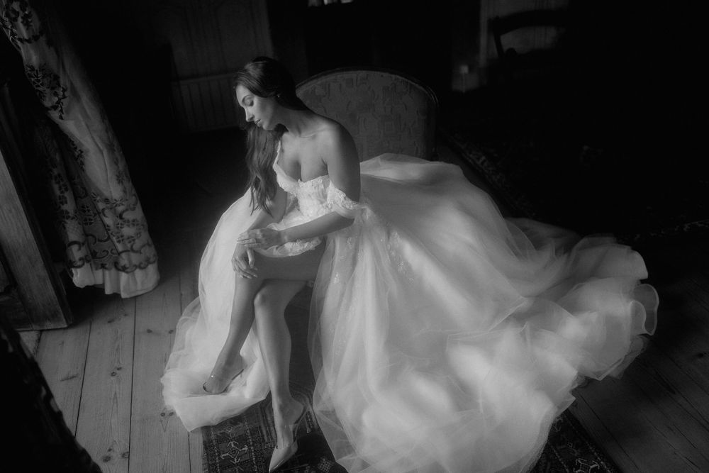 Black and white photo of a person in a flowing white wedding gown sitting pensively in a vintage interior setting.