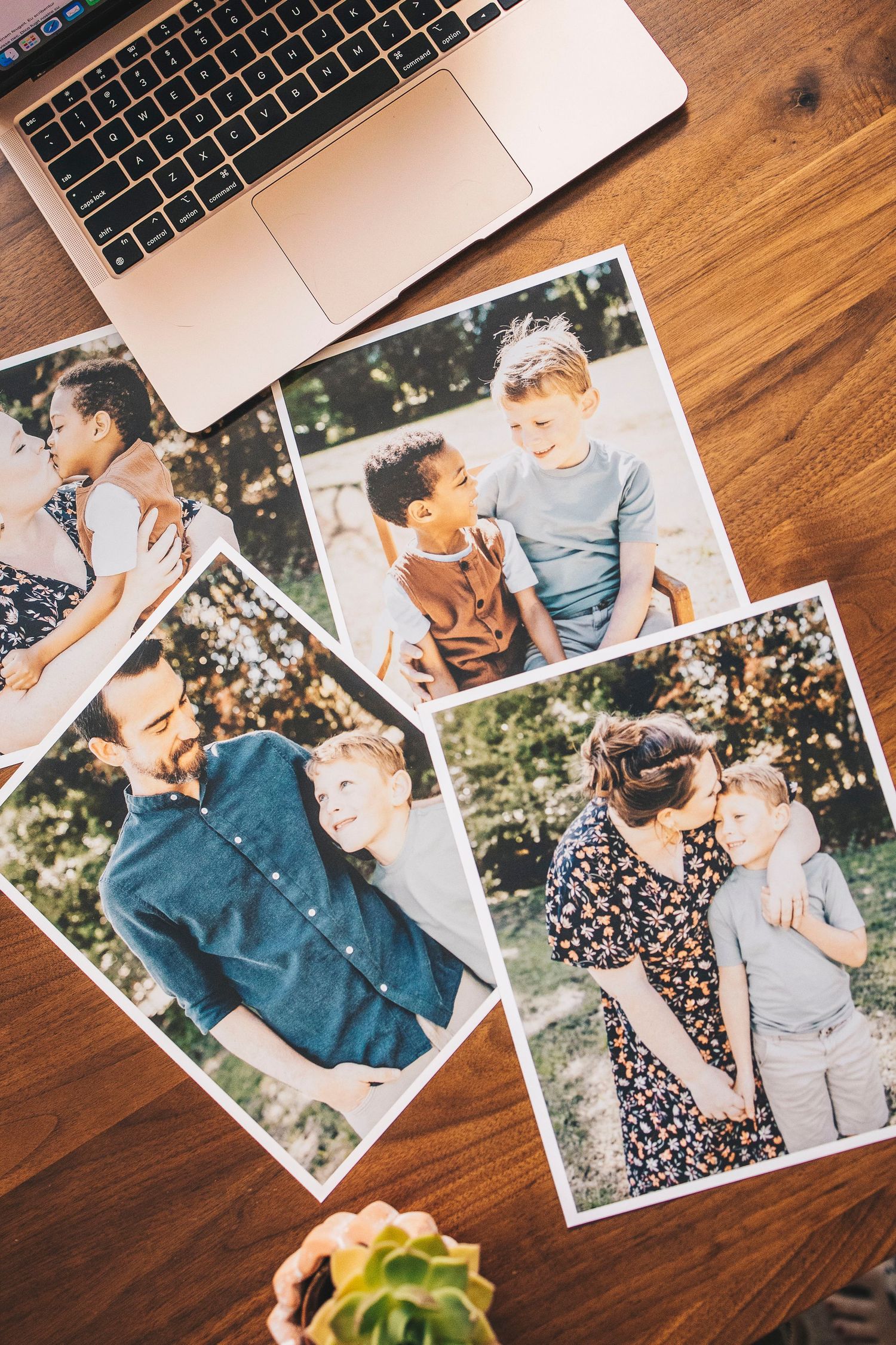 Printed family photos spread on a wooden desk beside a laptop and small succulent plant.